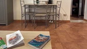 Una sala de estar con una mesa de comedor, sillas, un televisor en la pared, una planta pequeña y una revista en el suelo.