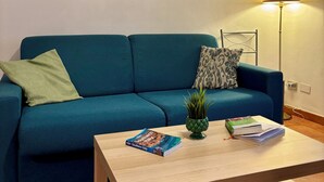 Una sala de estar con un sofá color esmeralda, una mesa de centro con libros y una planta, un cuadro de una escena costera y una lámpara de pie.