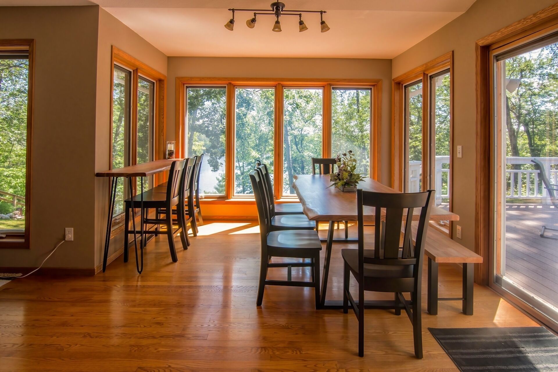 A dining area with a wooden table and chairs, large windows with a view of trees, and a ceiling light fixture.