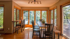 A dining area with a wooden table and chairs, large windows with a view of trees, and a ceiling light fixture. - Shady Crest. Pool table, fireplace, games, kayaks, Legos. 5 kings - 16ppl. (Brainerd)