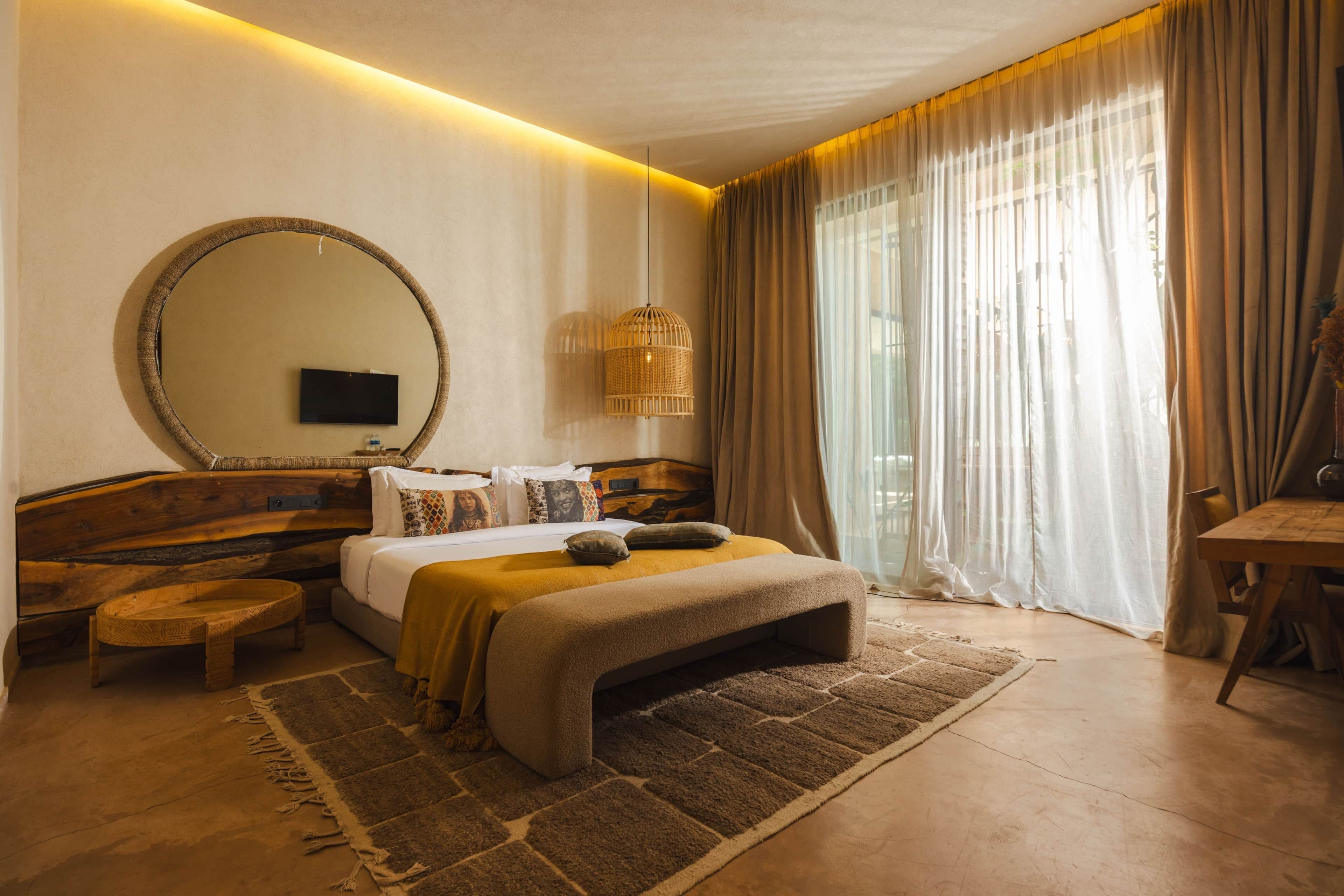 A bedroom with a large bed, a round mirror, a bedside table, a wooden bench, and a large window with curtains.