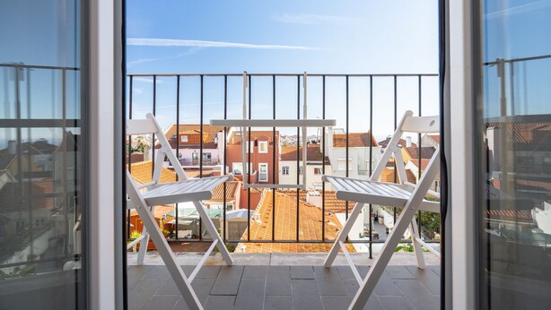 Un balcon avec des chaises blanches et une table, donnant sur un quartier résidentiel aux toits recouverts de tuiles rouges.