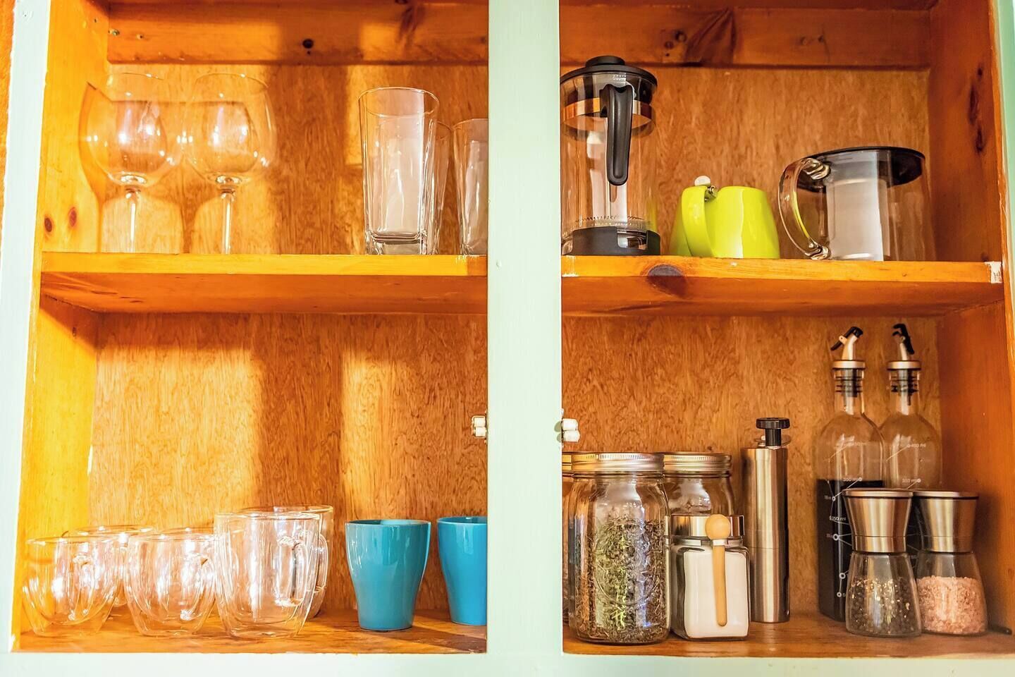 A wooden shelf with clear glasses, blue cups, a coffee maker, and various kitchen items.