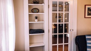 A room with a white door, a small shelf with decorative items, and a bed with a dark blanket. - Hudson Valley Apartment in a Historic Farmhouse w/ Sauna Near Stone Ridge Hamlet (Stone Ridge)