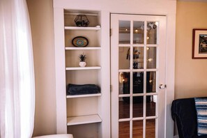 A room with a white door, a small shelf with decorative items, and a bed with a dark blanket. - Hudson Valley Apartment in a Historic Farmhouse w/ Sauna Near Stone Ridge Hamlet (Stone Ridge)