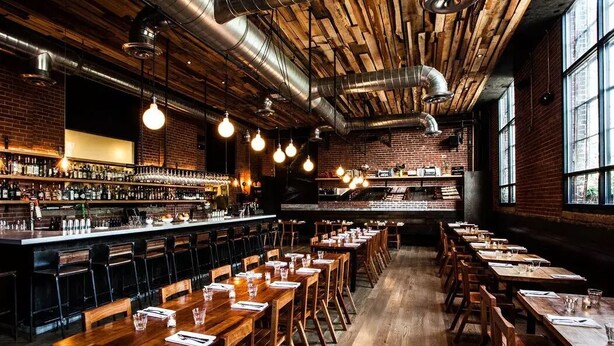 A restaurant with long wooden tables and chairs, a bar area with shelves of bottles, and a brick wall. - Abode Nashville - Germantown (Nashville)
