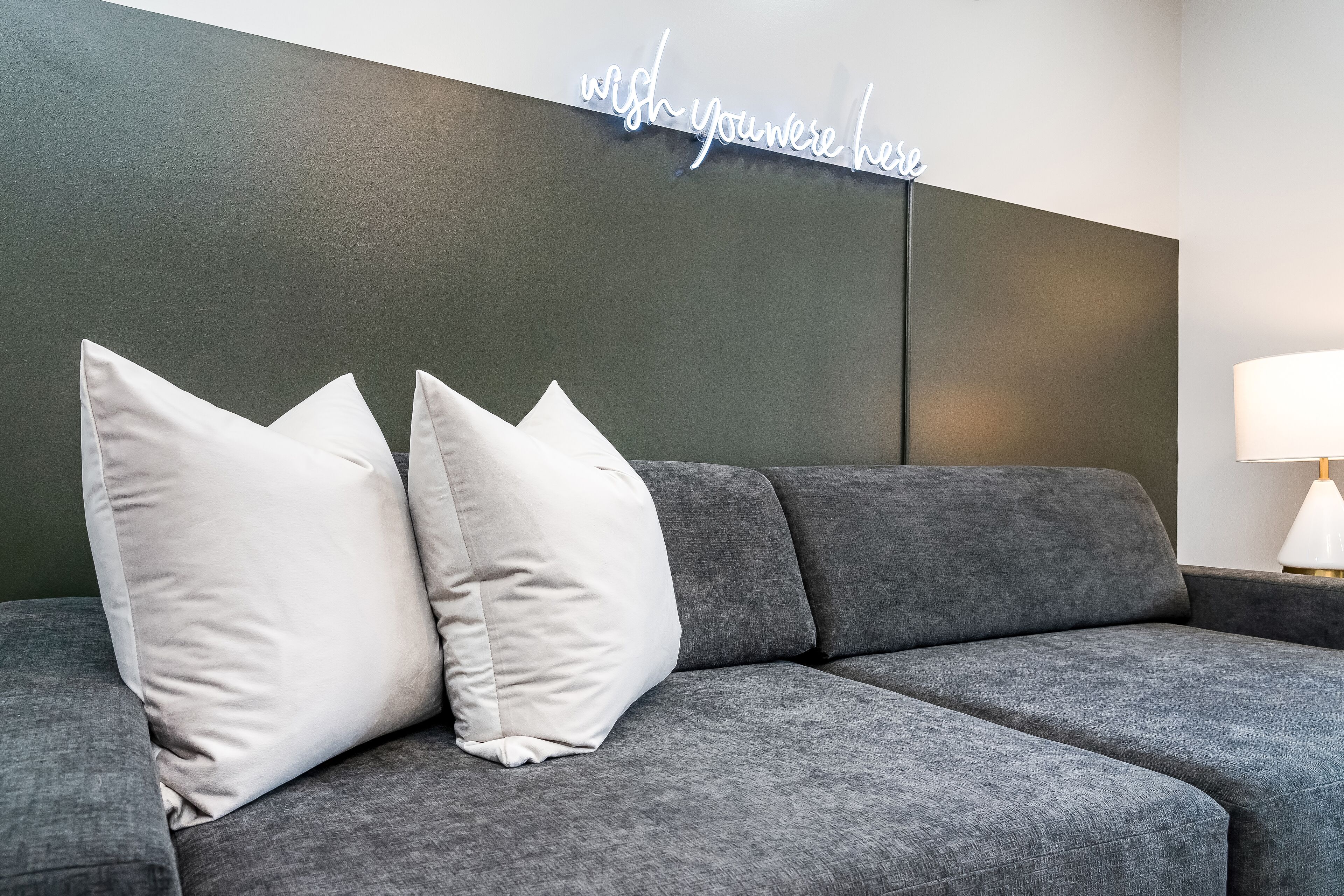 Un sofá gris con dos almohadas blancas y un letrero de neón que dice “wish you were here”.