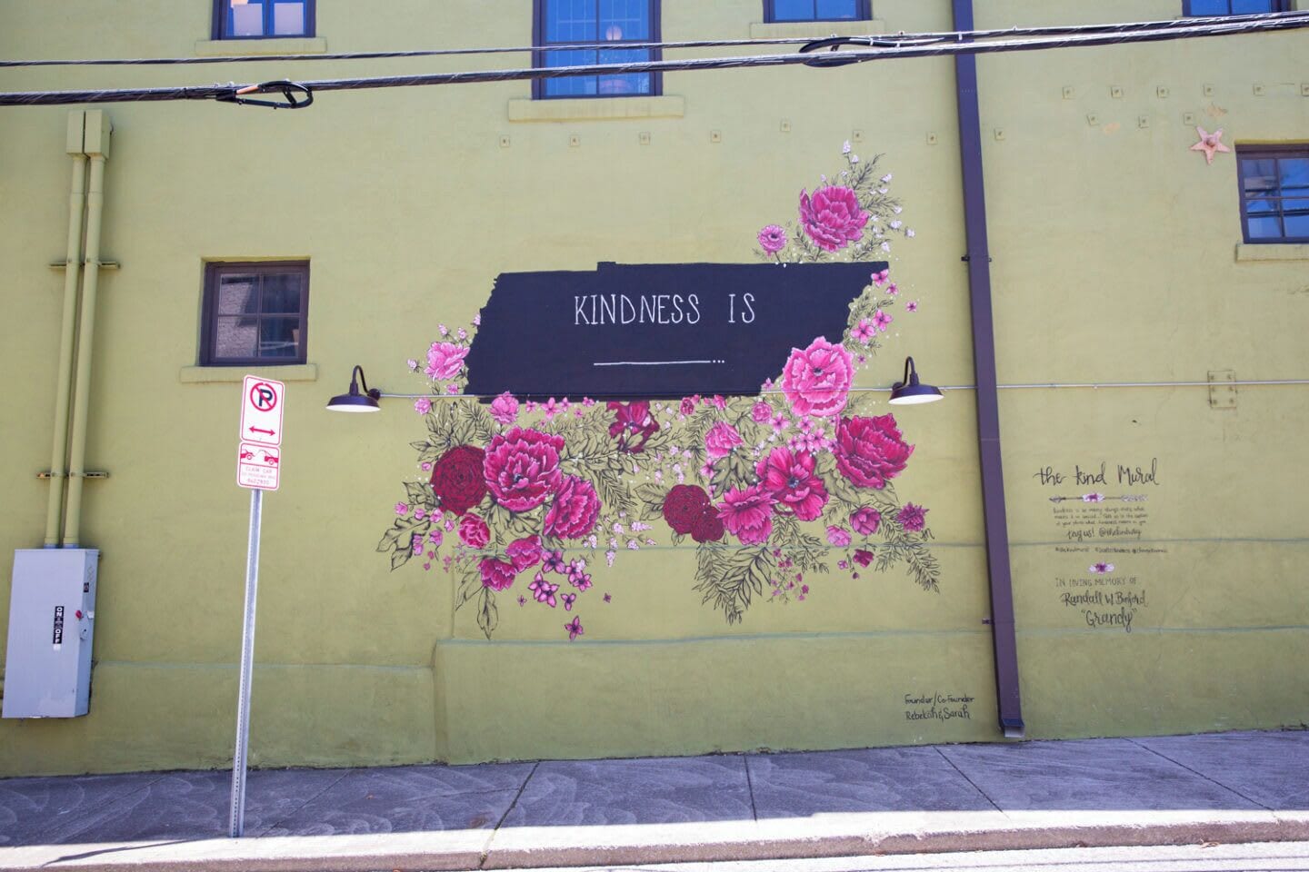 A mural with the text "KINDNESS IS" on a yellow wall, accompanied by floral artwork.