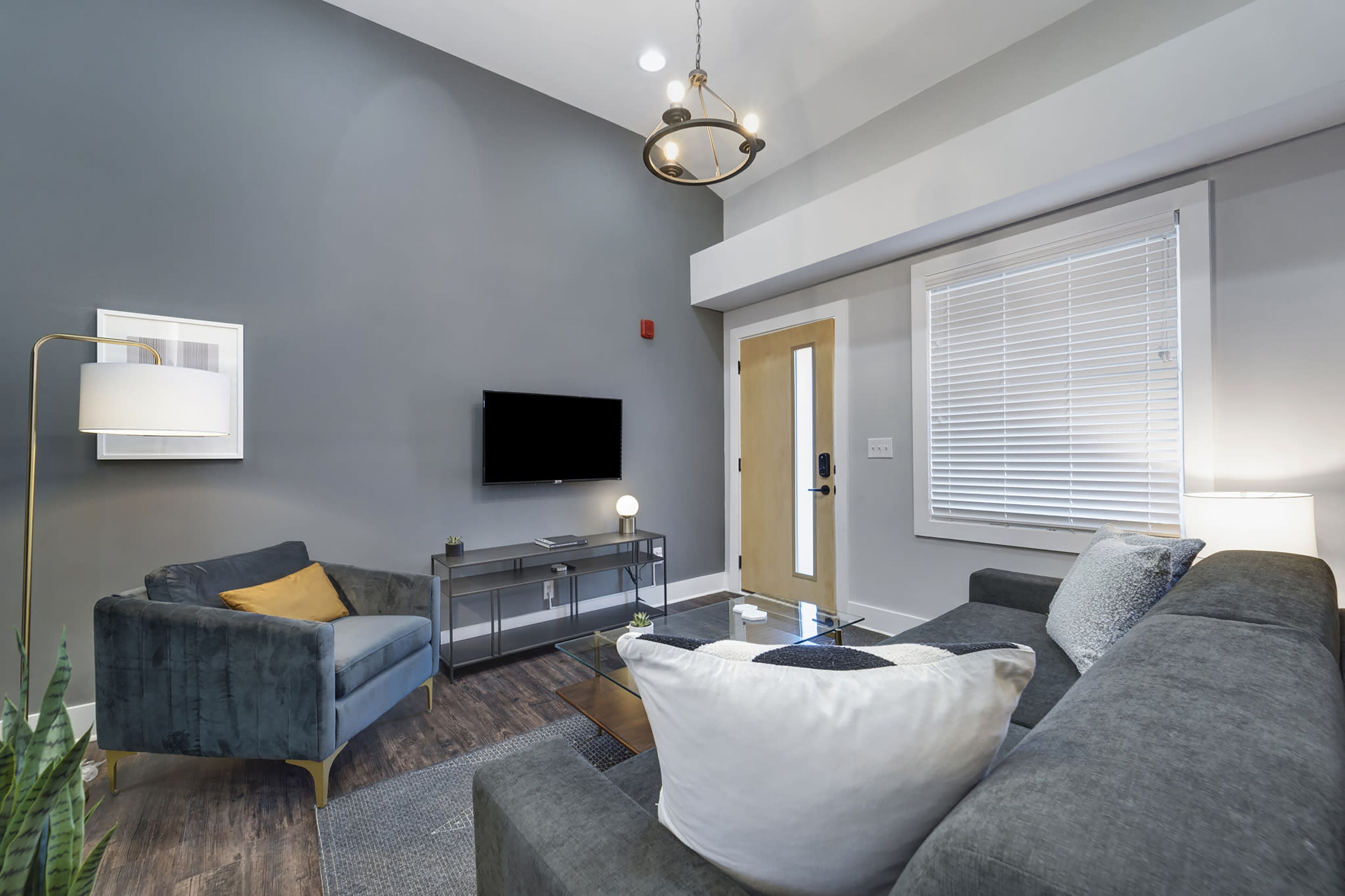 Una sala moderna con un sofá gris, un sillón azul, una mesa de centro de vidrio y un televisor de pantalla plana montado en la pared.