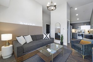 Una sala de estar moderna con un sofá gris, una mesa de centro de vidrio y una cocina al fondo.