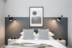 Un dormitorio moderno con una cama, dos mesitas de noche, una obra de arte enmarcada y dos lámparas de pie.