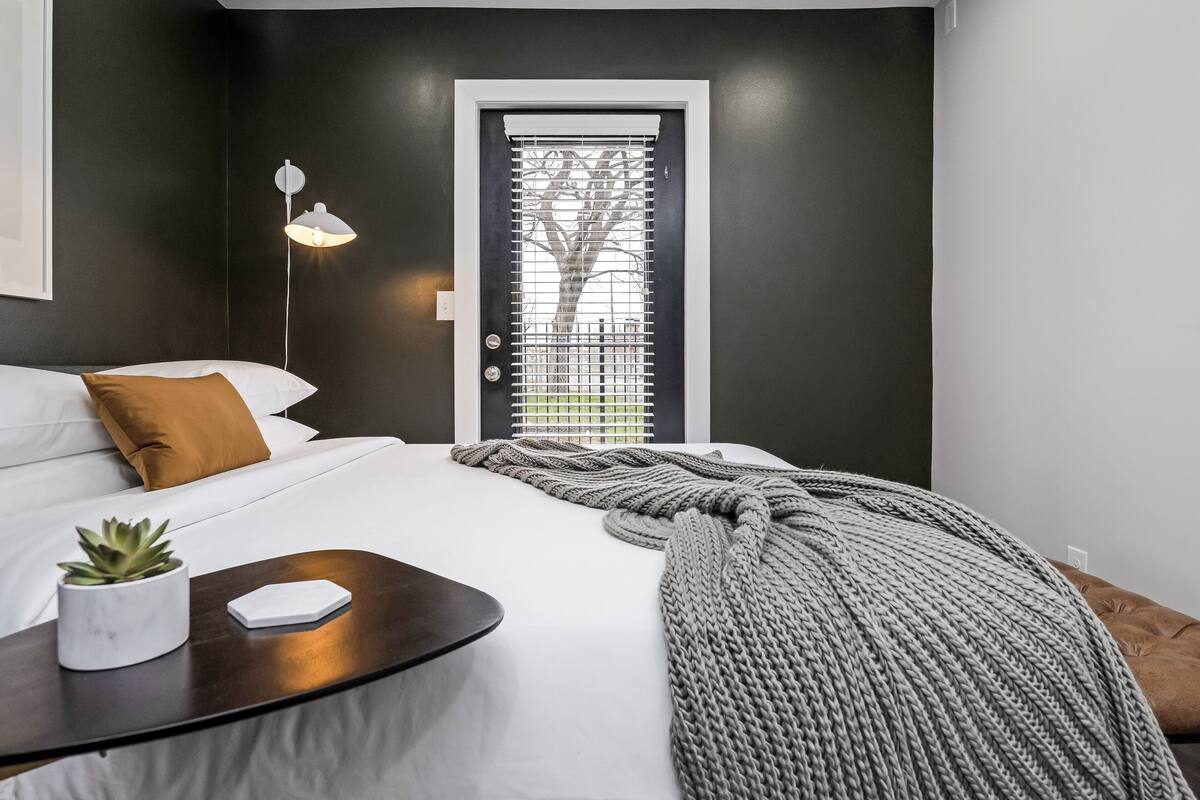 A modern hotel room with a large bed, a bedside table with a lamp, a potted plant, and a view through a window with blinds.