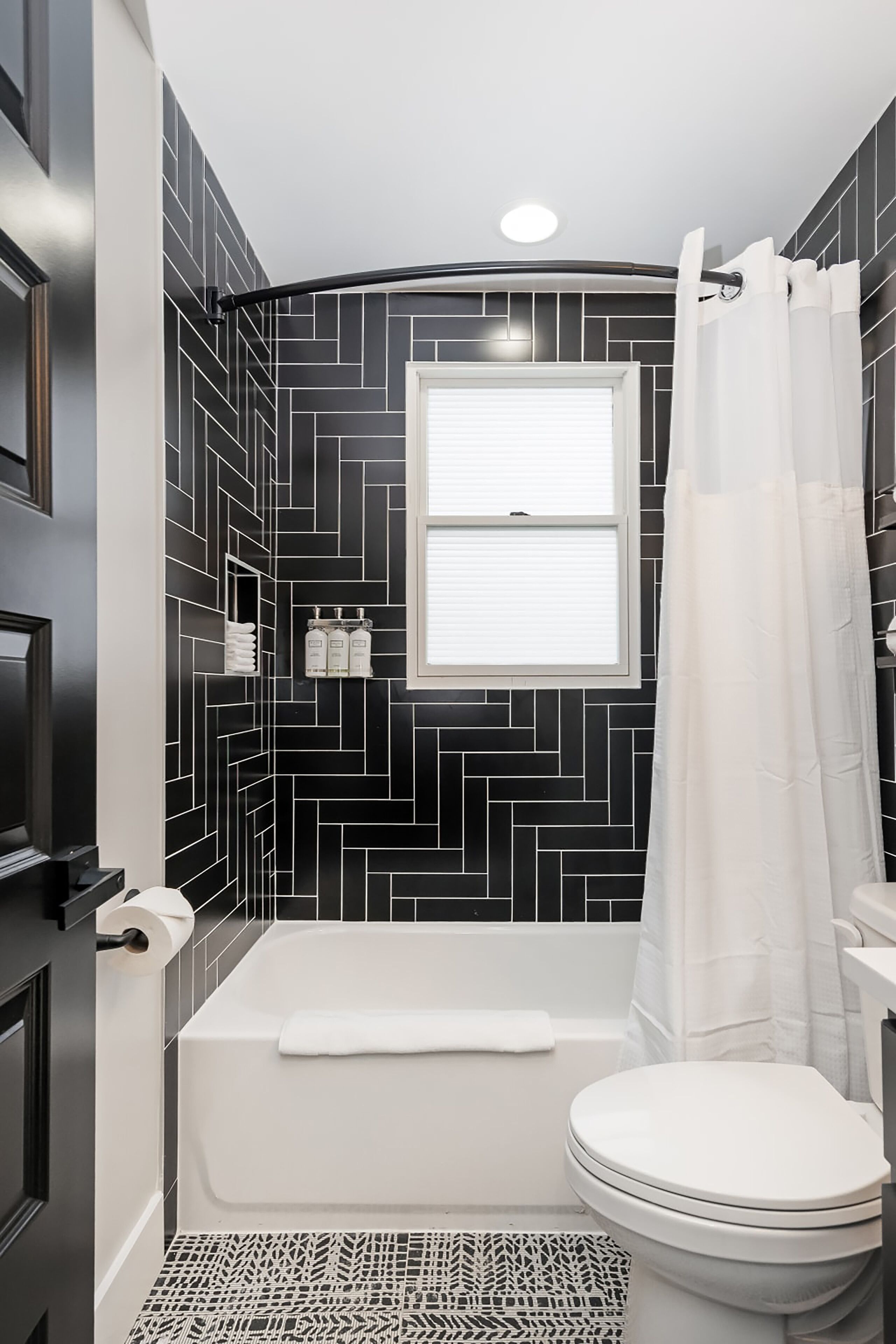 Un baño con salpicadero de azulejos blancos y negros en patrón de espiga, una bañera blanca, un inodoro blanco y una ventana con cortina blanca.
