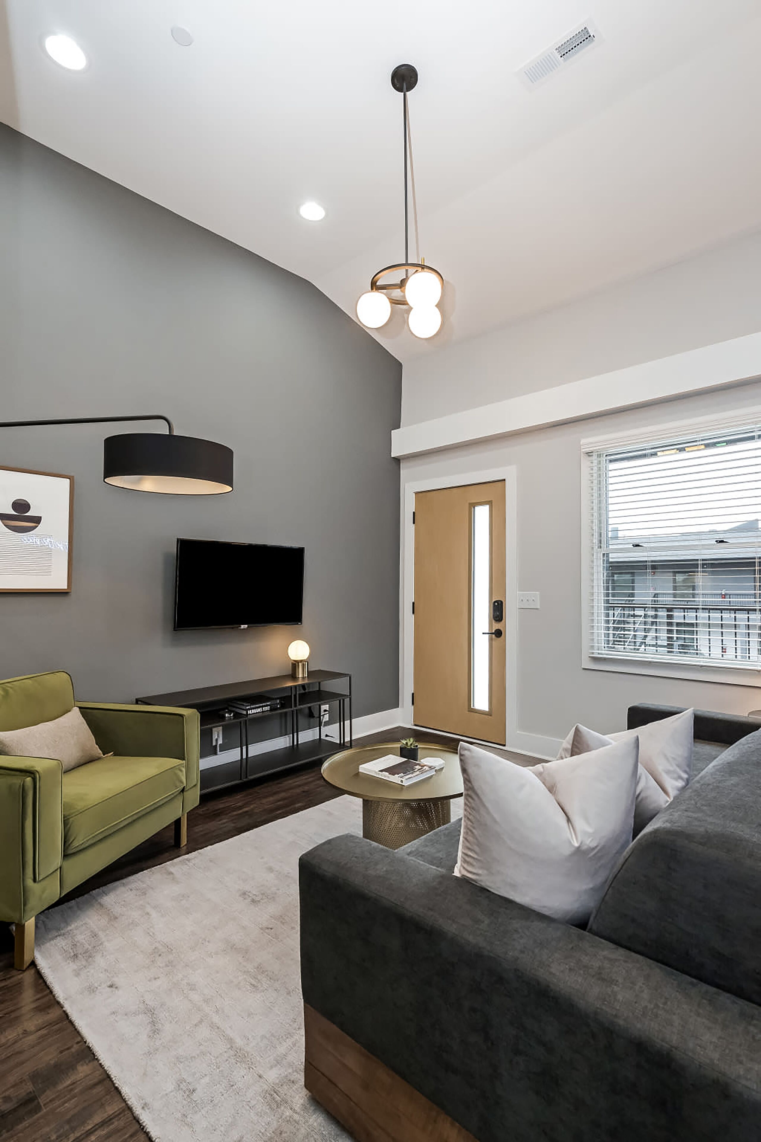 Un salon moderne avec un canapé gris, un fauteuil vert, un téléviseur à écran plat et un luminaire suspendu au plafond.
