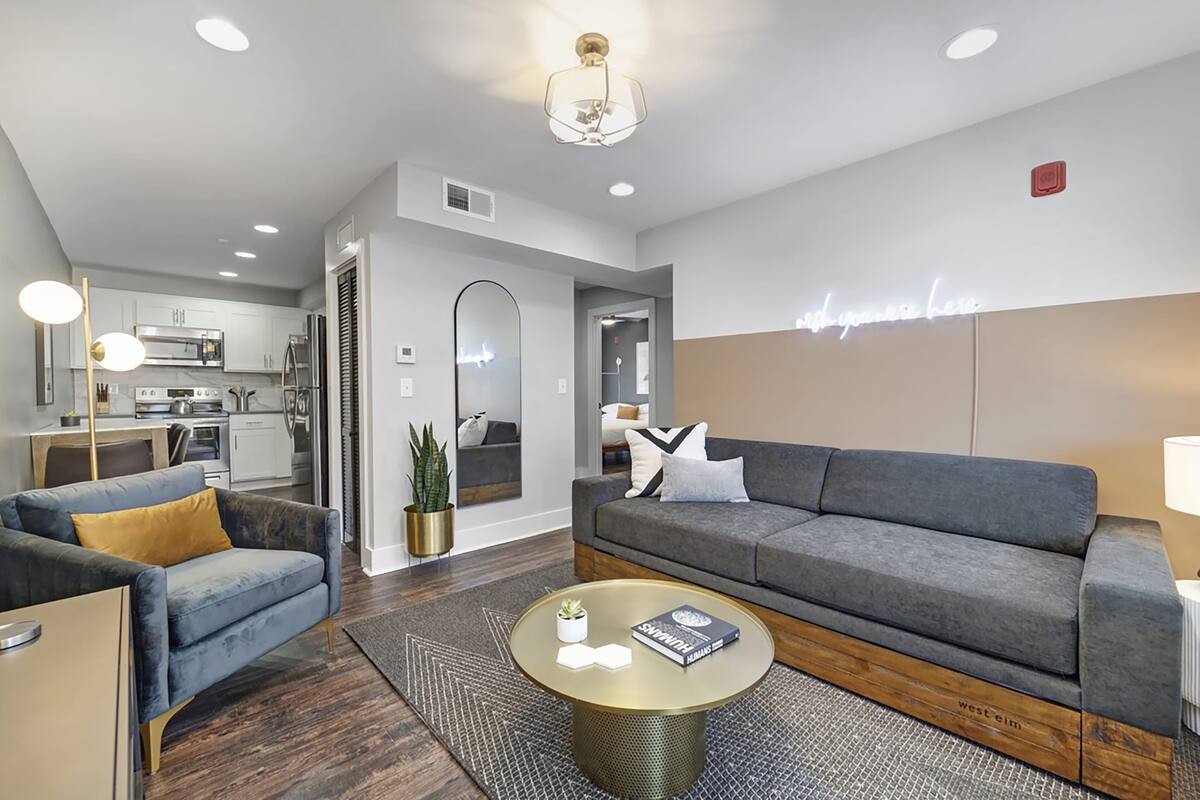 A modern living room with a grey sofa, a blue armchair, and a coffee table. There is a kitchen in the background with stainless steel appliances.