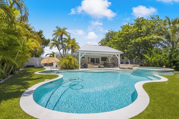 A swimming pool with a white deck, surrounded by palm trees and a covered patio area.