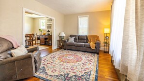 A living room with a brown leather sofa, a patterned rug, and a dining area visible through an open doorway.