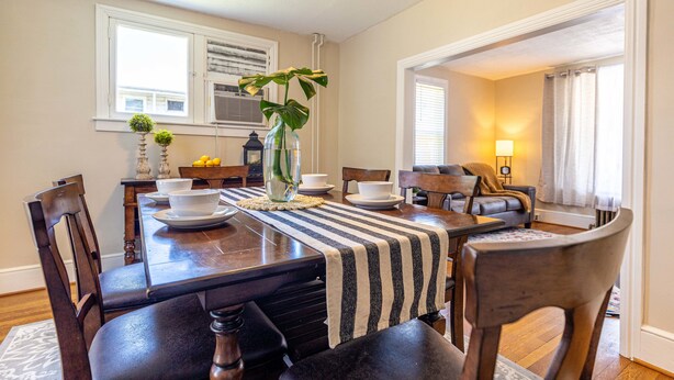 A dining room with a wooden table, chairs, and a striped table runner. There is a vase with a plant on the table.