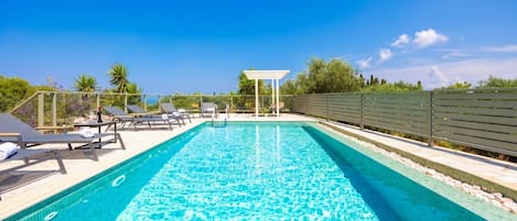 A rectangular swimming pool with clear blue water, surrounded by lounge chairs and a pergola structure.