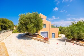 A two-story building with a garage, surrounded by a stone-paved area and trees. - Villa Canova - Three Bedroom Villa, Sleeps 6 (Ionian Islands)