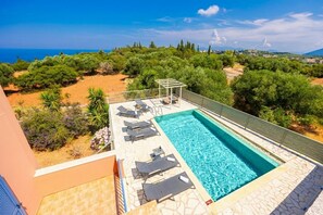 A rooftop swimming pool with lounge chairs and a pergola, surrounded by greenery and a view of the sea.