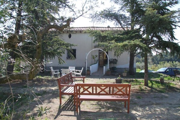 A rustic building with a white facade and a tiled roof, surrounded by trees and a garden.