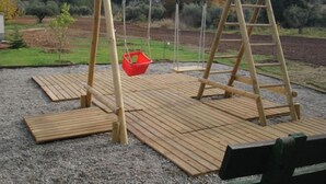 Un ensemble de balançoires en bois, avec un panier rouge suspendu à l’une des balançoires, installé sur une terrasse en bois dont le sol est recouvert de gravier.