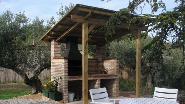 Un espace barbecue extérieur avec un gril en briques, une pergola en bois et des chaises en plastique blanches.