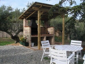 Ein Außenbereich mit Grillplatz, Ziegelgrill, einer Holzpergola und weißen Kunststoffstühlen.