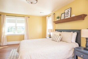 Quarto com cama, mesas de cabeceira, janela com cortinas, luminária no teto e prateleira com objetos decorativos.