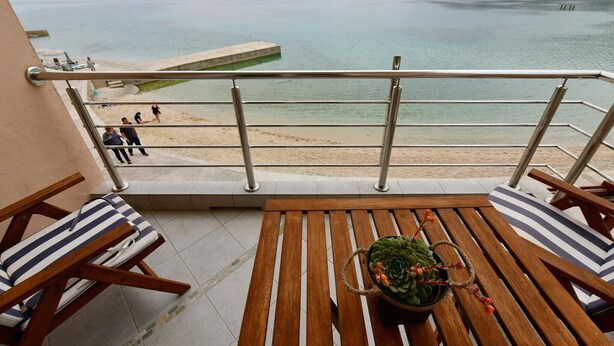 Un balcon agrémenté de meubles en bois et d’une plante en pot, offrant une vue sur une plage et une mer calme, avec des montagnes en arrière-plan.