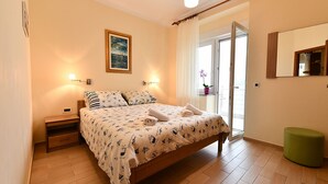 A bedroom with a bed, bedside tables, a nightstand, a green ottoman, a door to a balcony, and a painting on the wall. - Holiday apartment with balcony and sea view (Baška)
