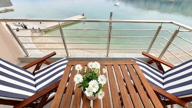 Un balcone con un tavolo e sedie in legno, un vaso di fiori e vista su un lago calmo con barche.