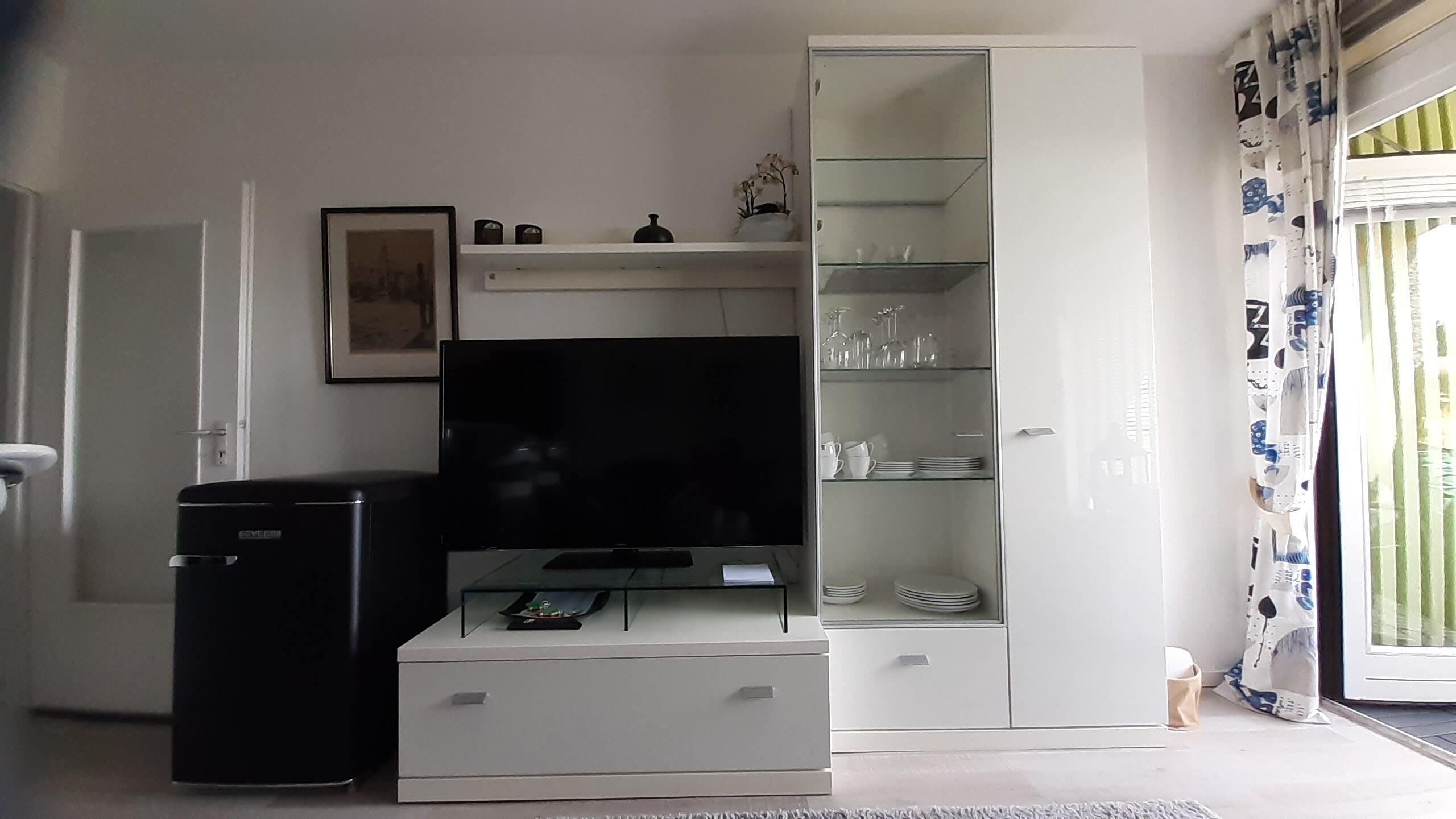 Een woonkamer met een flatscreen televisie, een zwarte koelkast, een wit kastje met planken en een deur naar een balkon.