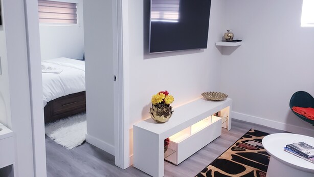 Et moderne hotelværelse med seng, fjernsyn ophængt på væggen, et hvidt konsolbord med blomsterstilleben og et rundt, hvidt sofabord samt en gulvtæppe med mønster.