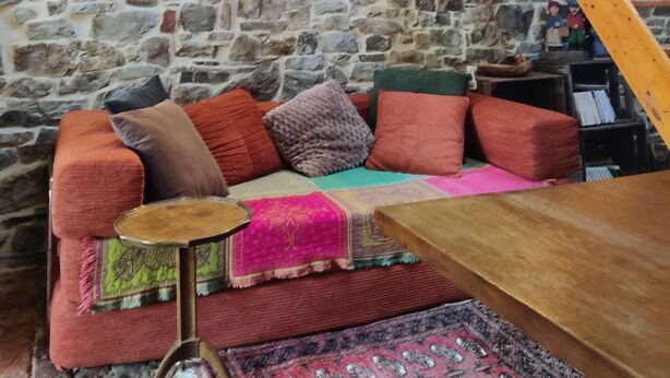 A rustic room with a stone wall, a wooden staircase, a colorful sofa, and a painting. - La Grange- Historic flair and modern comfort in the midst of nature (Sourbrodt, Waimes (Belgien))