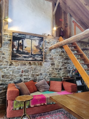 A rustic room with a stone wall, a wooden staircase, a colorful sofa, and a painting.