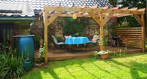 A wooden pergola with a table and chairs, a stone wall, and a blue barrel in a garden setting.