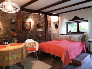 A cozy bedroom with a stone wall, a wooden ceiling, a bed with a red blanket, a chair, a small table with bottles, and a window with a view of greenery.