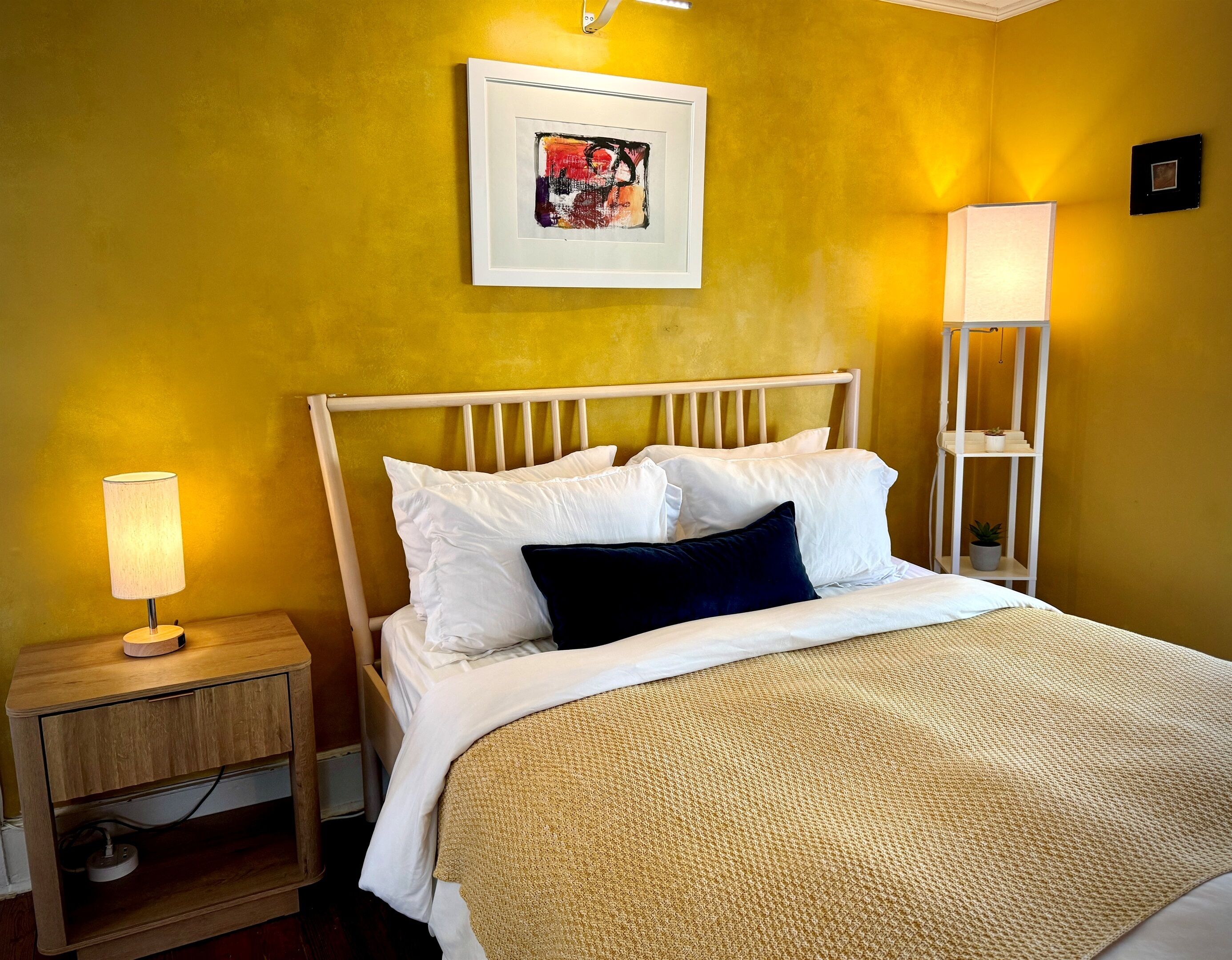 Una cama con ropa de cama blanca, un almohadón oscuro y una manta texturizada. Hay una mesita de noche con una lámpara y un pequeño estante con una planta. Un cuadro enmarcado cuelga de la pared sobre la cama.