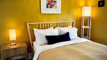 Una cama con ropa de cama blanca, un almohadón oscuro y una manta texturizada. Hay una mesita de noche con una lámpara y un pequeño estante con una planta. Un cuadro enmarcado cuelga de la pared sobre la cama.