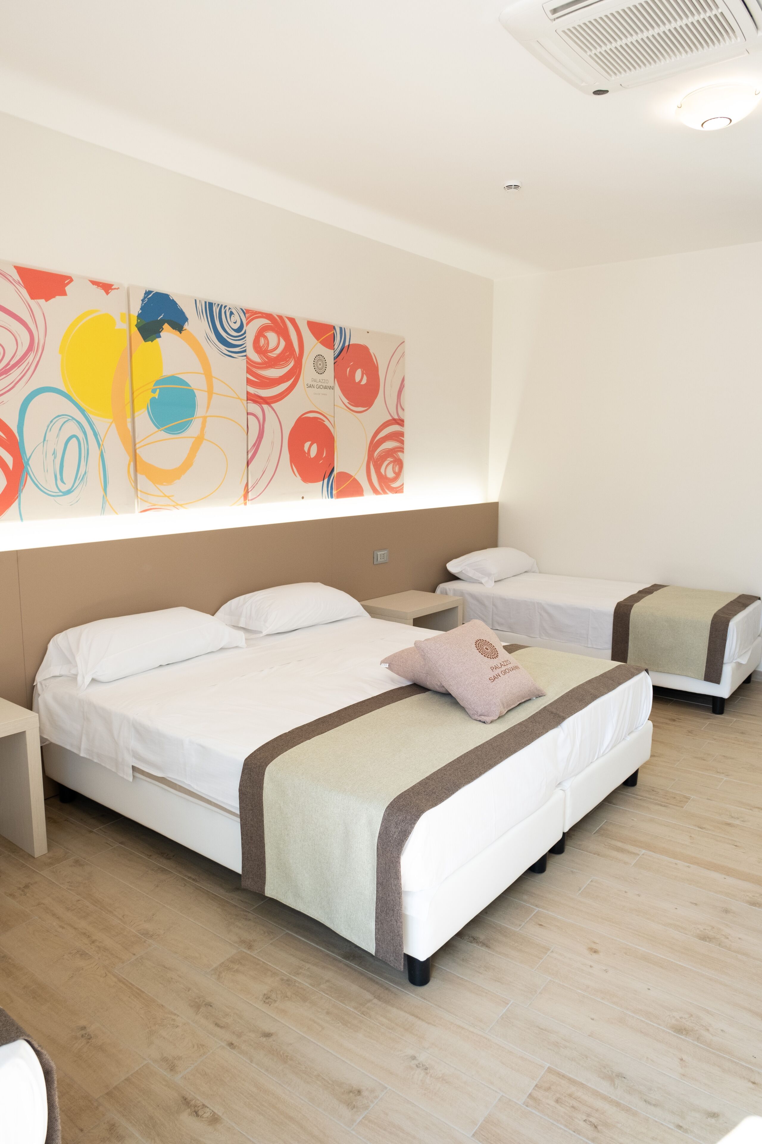 A hotel room with two beds, a large abstract painting on the wall, and a ceiling vent.