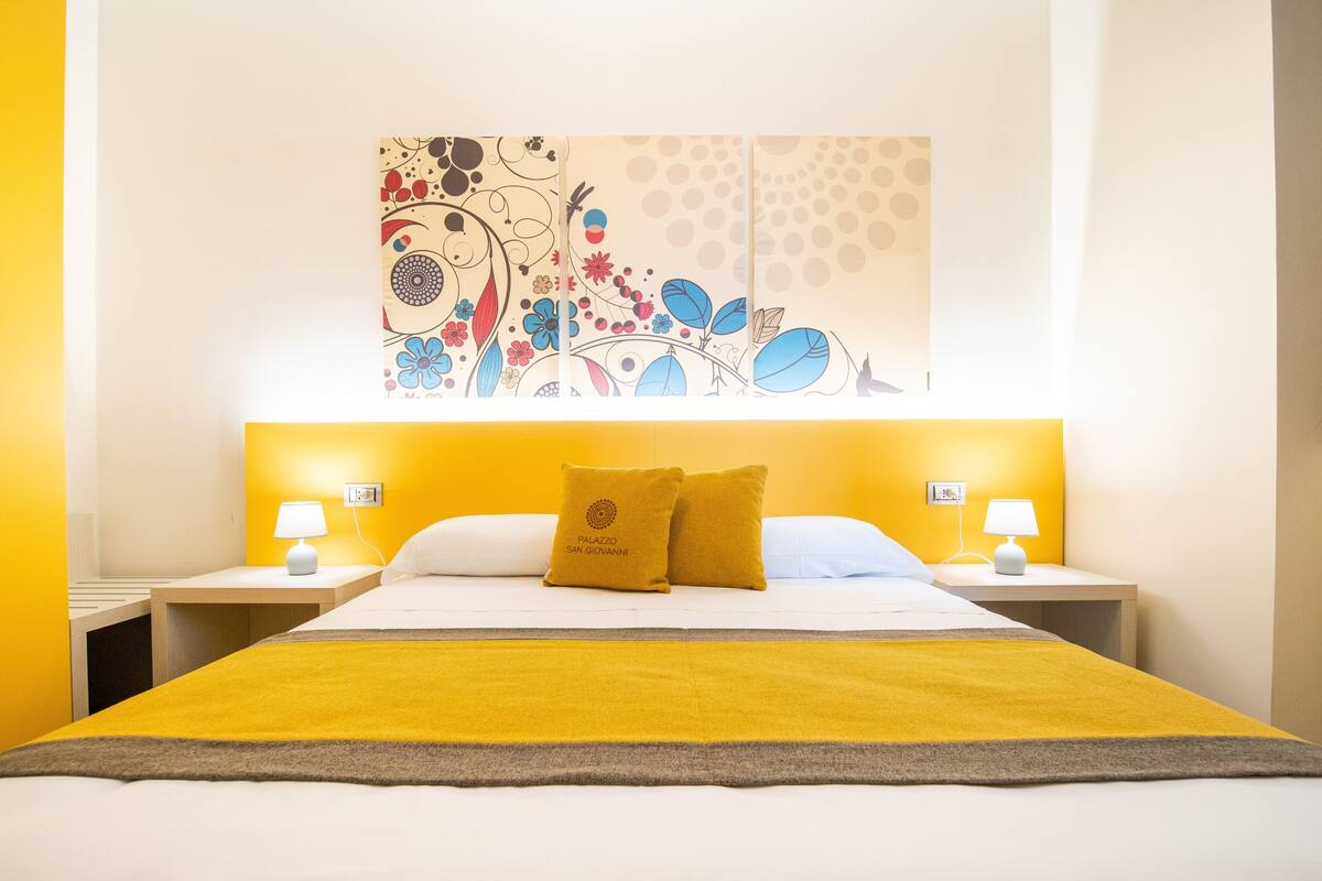 Een hotelkamer met een bed, twee nachtkastjes, een kunstwerk aan de muur en een gele accentmuur.