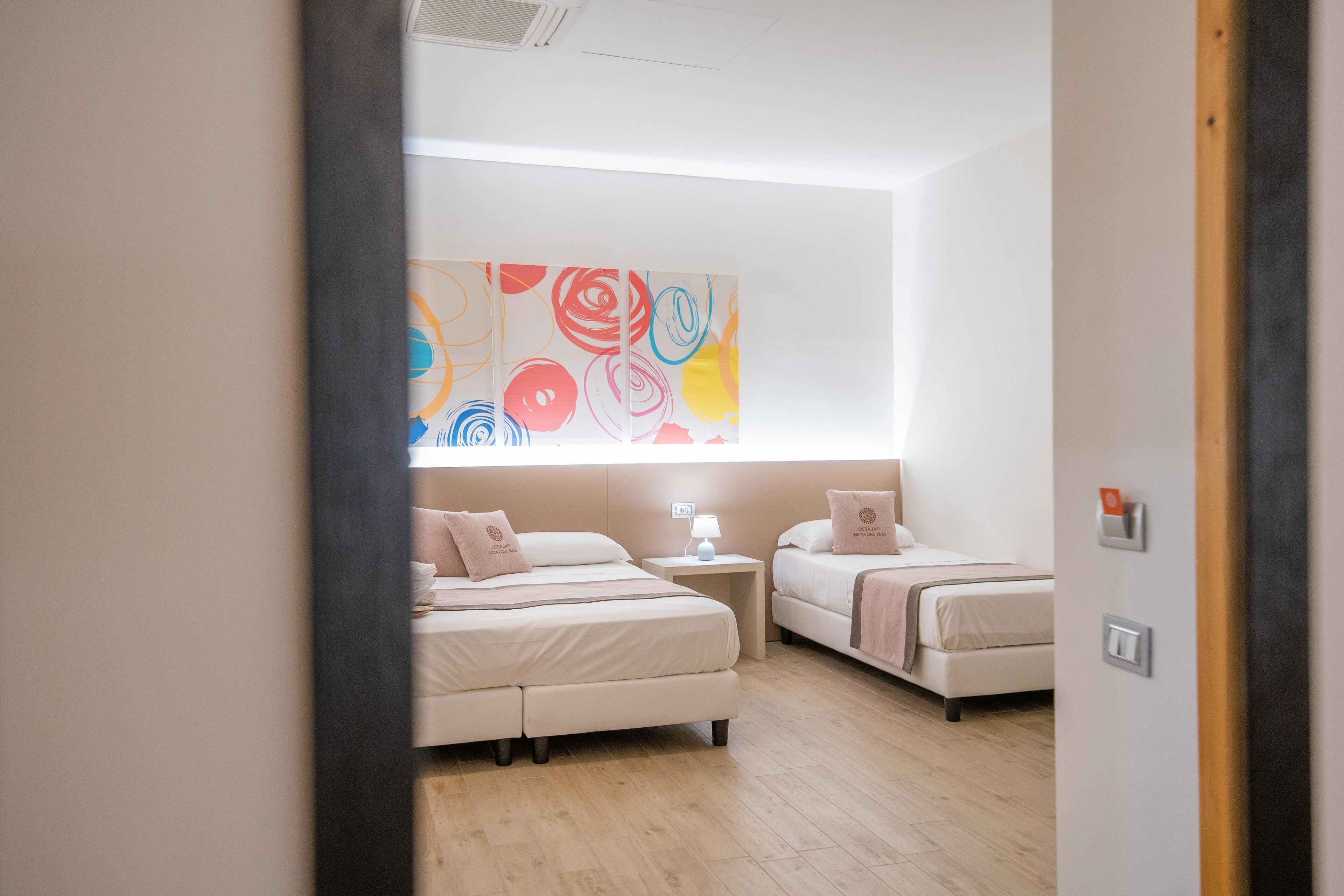 Une chambre d’hôtel moderne avec deux lits, une grande peinture abstraite au mur, une table de chevet avec une lampe et un plancher en bois.