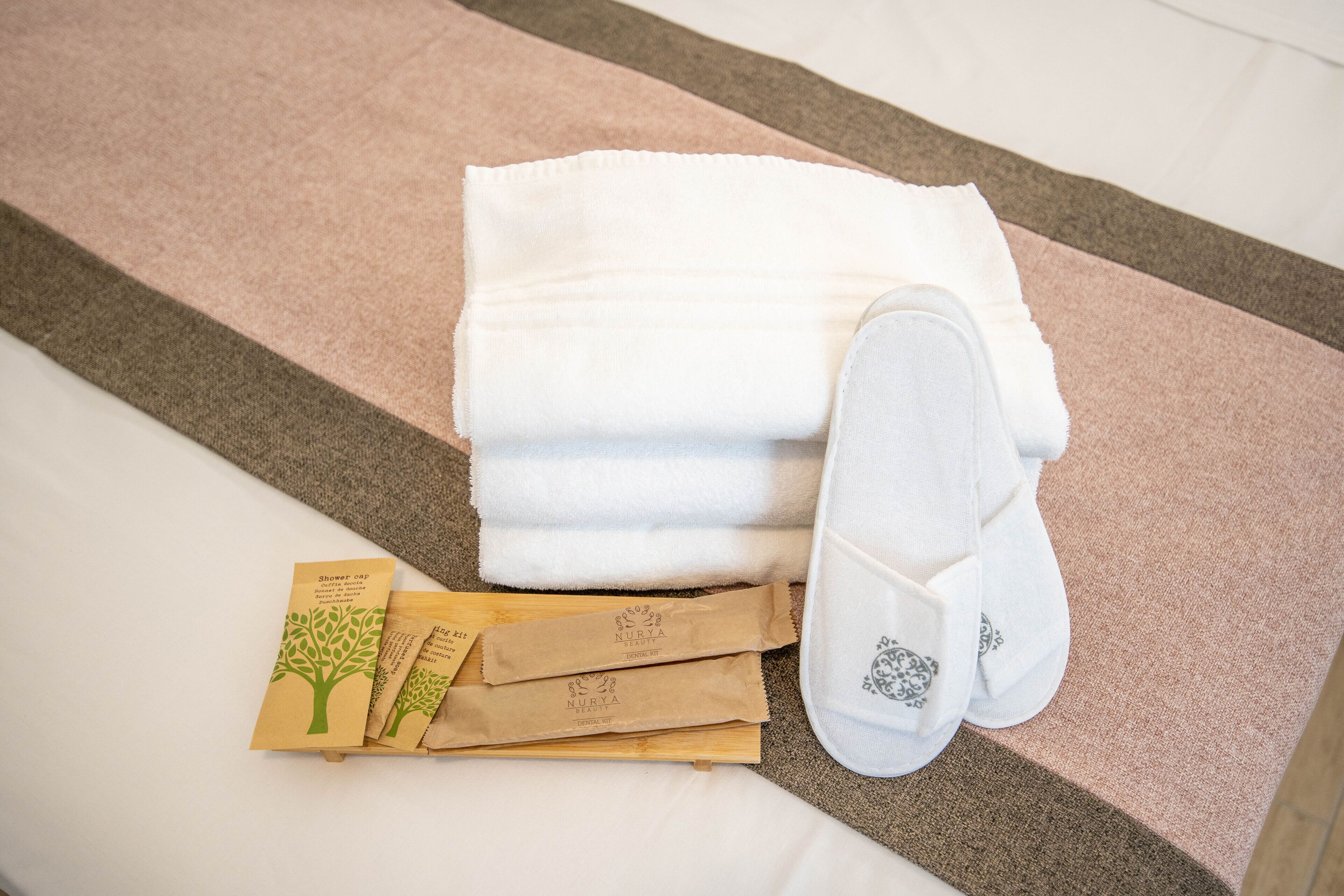 Un lit bien fait, avec une serviette blanche et des chaussons, un tapis de bain marron et deux bonnets de douche, le tout posé sur une surface en bois.