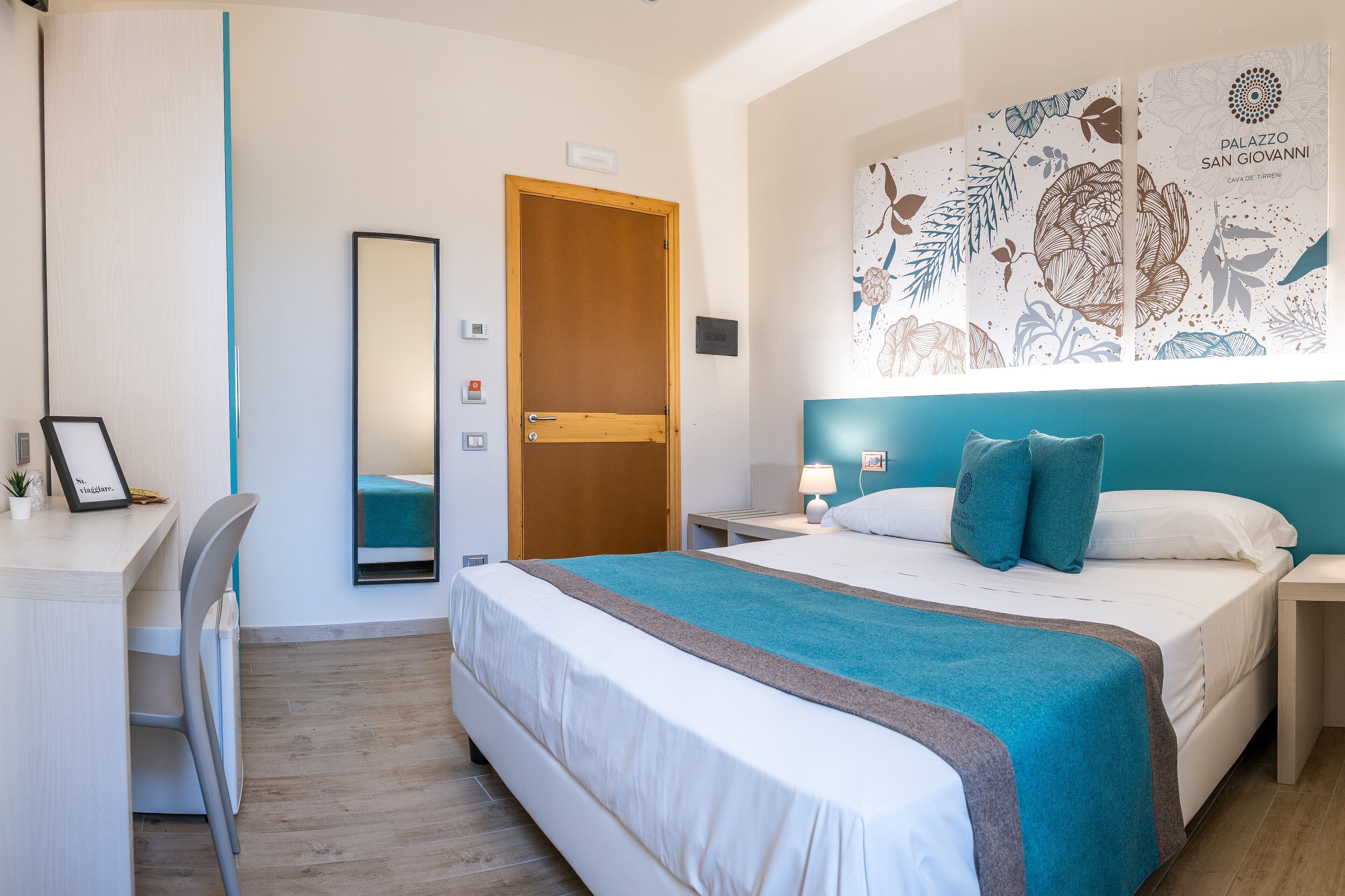 A hotel room with a bed, a desk, a chair, a mirror, a nightstand, and a wall with a floral pattern.