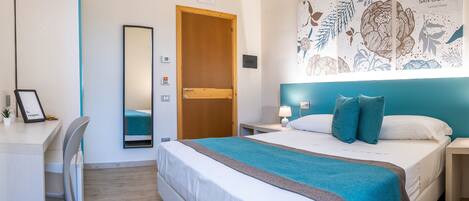 A hotel room with a bed, a desk, a chair, a mirror, a nightstand, and a wall with a floral pattern.