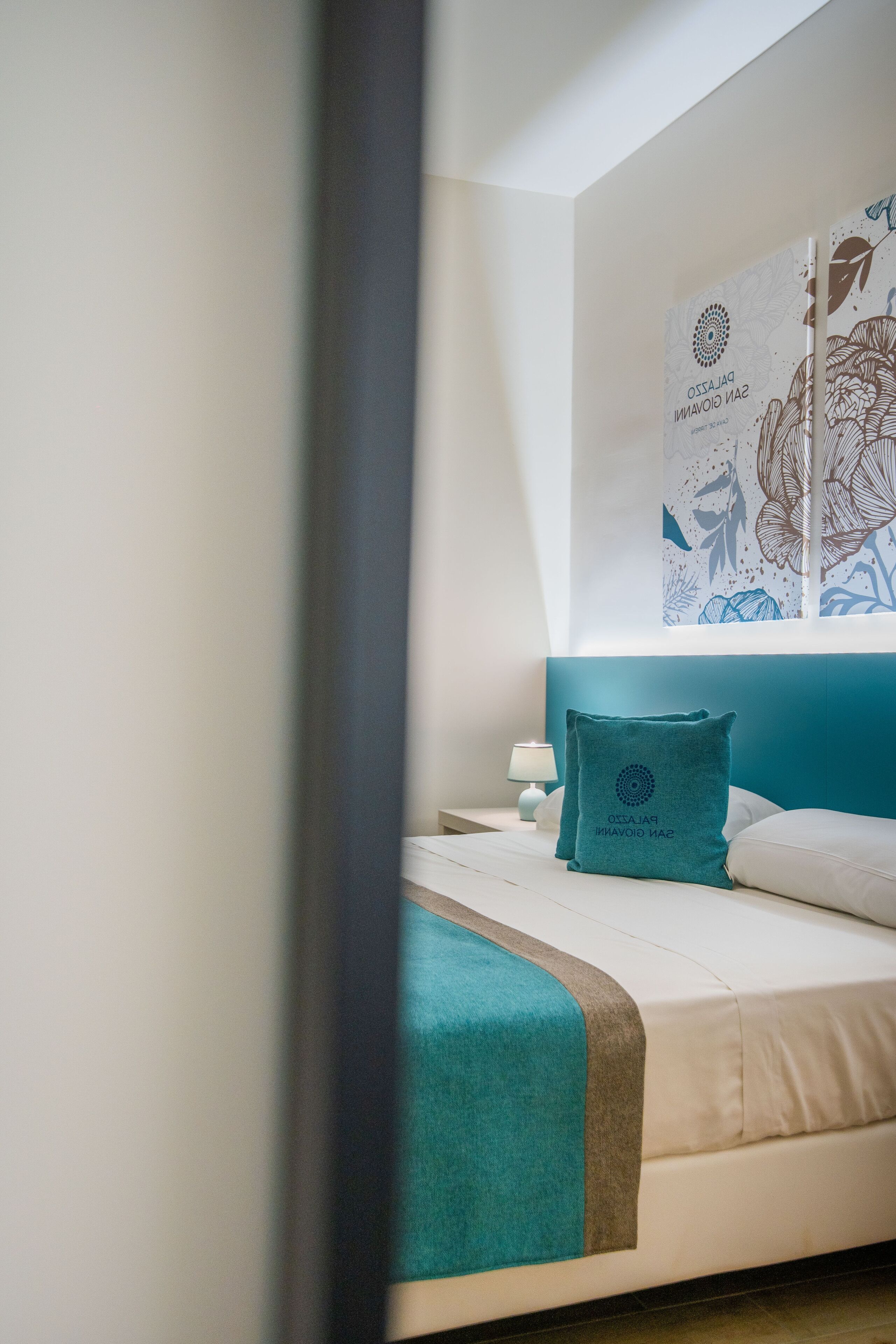 Ein Schlafzimmer mit einem Bett, einem türkisfarbenen Kissen, einem weißen Kissen, einem Nachttisch und einer Wand mit floralem Dekor.