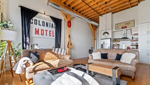 Ein gemütliches Wohnzimmer mit zwei Sofas, einem Couchtisch und einem Esstisch. Eine Gitarre hängt an der Wand, und es ist ein Schild mit der Aufschrift "Colonial Motel" zu sehen.