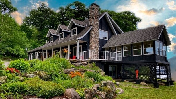 A large, dark-colored house with a stone chimney, multiple windows, and a porch. - Stillwater Lakehouse • 20 Acre Lakefront • Sandy Beach • Dock • Firepit! (Port Carling)